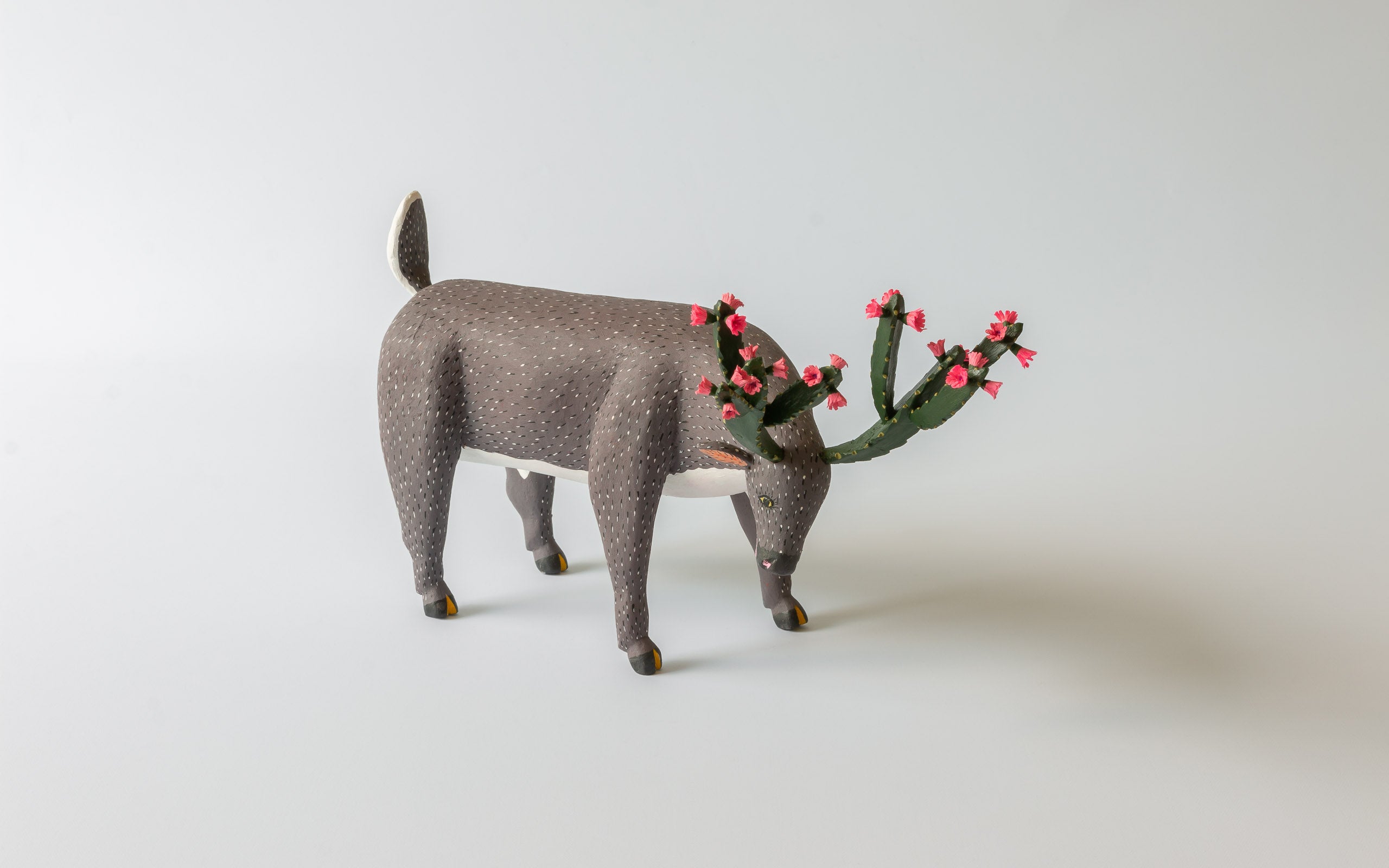 Gräsender Hirsch mit Geweih aus Kakteen mit rosa Blüten, braunes Fell.